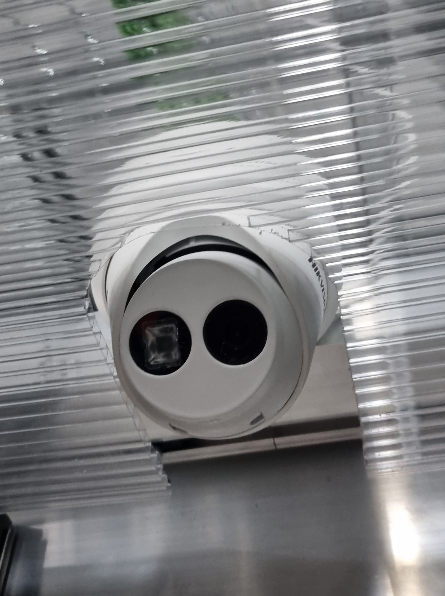 Biała kamera kopułkowa Hikvision zamontowana pod transparentnym, żebrowanym dachem z tworzywa sztucznego, widok z dołu.