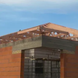 Surowy stan budowy domu z czerwonej cegły z widoczną drewnianą konstrukcją dachu w trakcie montażu, na tle błękitnego nieba.