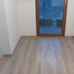 Nowo ułożone panele podłogowe w jasnym odcieniu drewna, widoczne przy ścianie z szarym akcentem i gniazdkami elektrycznymi oraz brązowymi drzwiami balkonowymi z widokiem na zewnątrz i grzejnikiem.