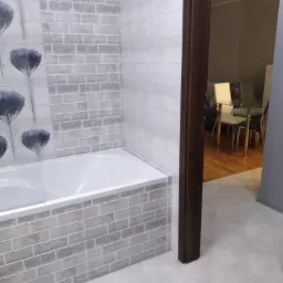 Wykończona łazienka z wanną obłożoną szarymi płytkami imitującymi cegłę, ściana z motywem kwiatowym, widok na salon przez otwarte drzwi.