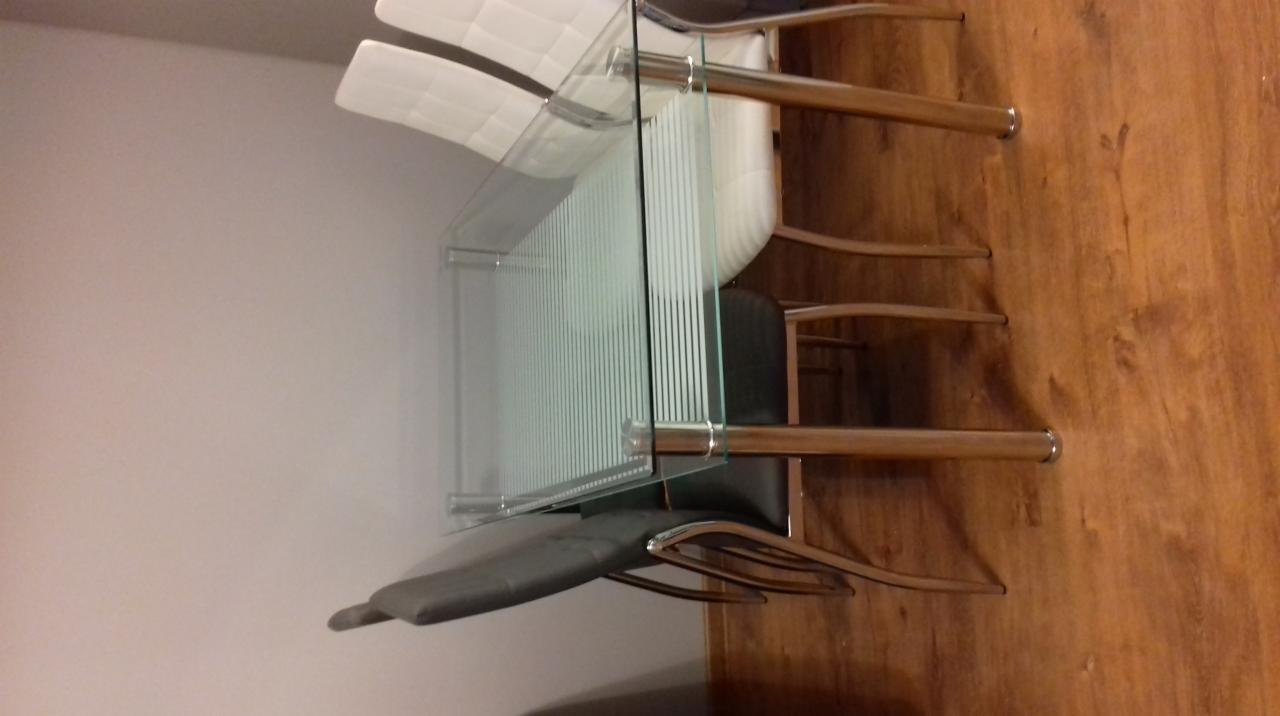 Szklany stół z chromowanymi nogami i krzesłami z białymi i szarymi siedziskami na tle drewnianej podłogi i jasnej ściany.