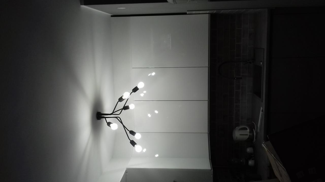 Nowoczesna łazienka z czarną armaturą, szarą cegłą na ścianie i oryginalną lampą z żarówkami na tle białych szafek.