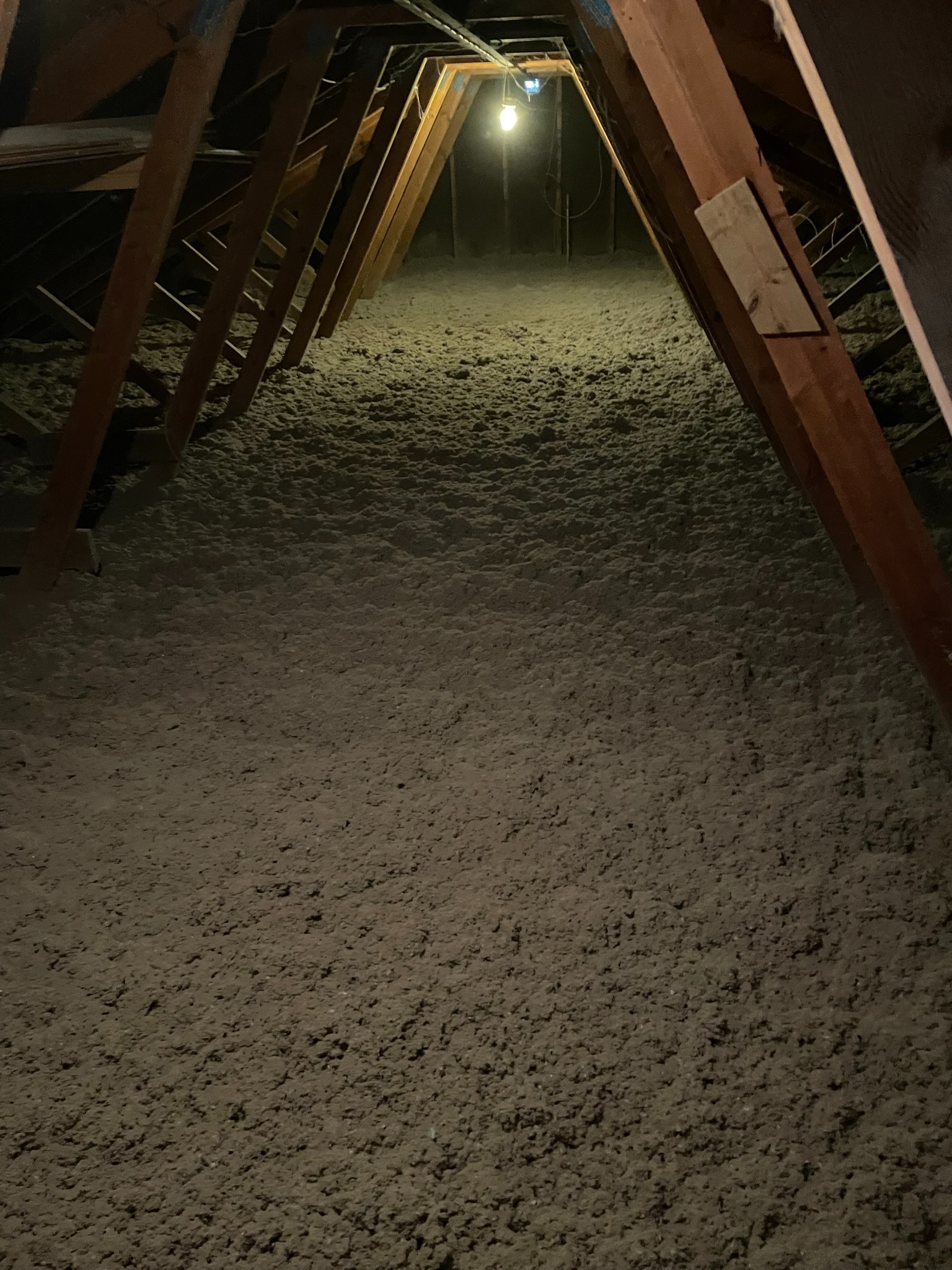 Poddasze ocieplone szarą wełną mineralną, widoczne drewniane krokwie i słabe oświetlenie żarówką