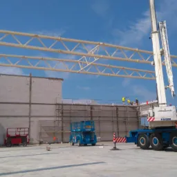 Wznoszenie konstrukcji hali: stalowe belki stropowe i ściany z bloczków, widoczny dźwig Faun i podnośniki koszowe na placu budowy.