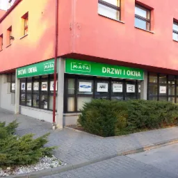 Fasada budynku z szyldem 'M-ATA DRZWI I OKNA', prezentująca ekspozycję okien i drzwi różnych producentów, z widocznymi logotypami marek na szybach.