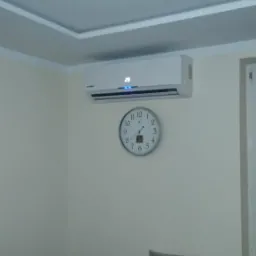 Biała jednostka wewnętrzna zamontowana na ścianie nad okrągłym zegarem w salonie z szarą sofą i dekoracyjną listwą sufitową.