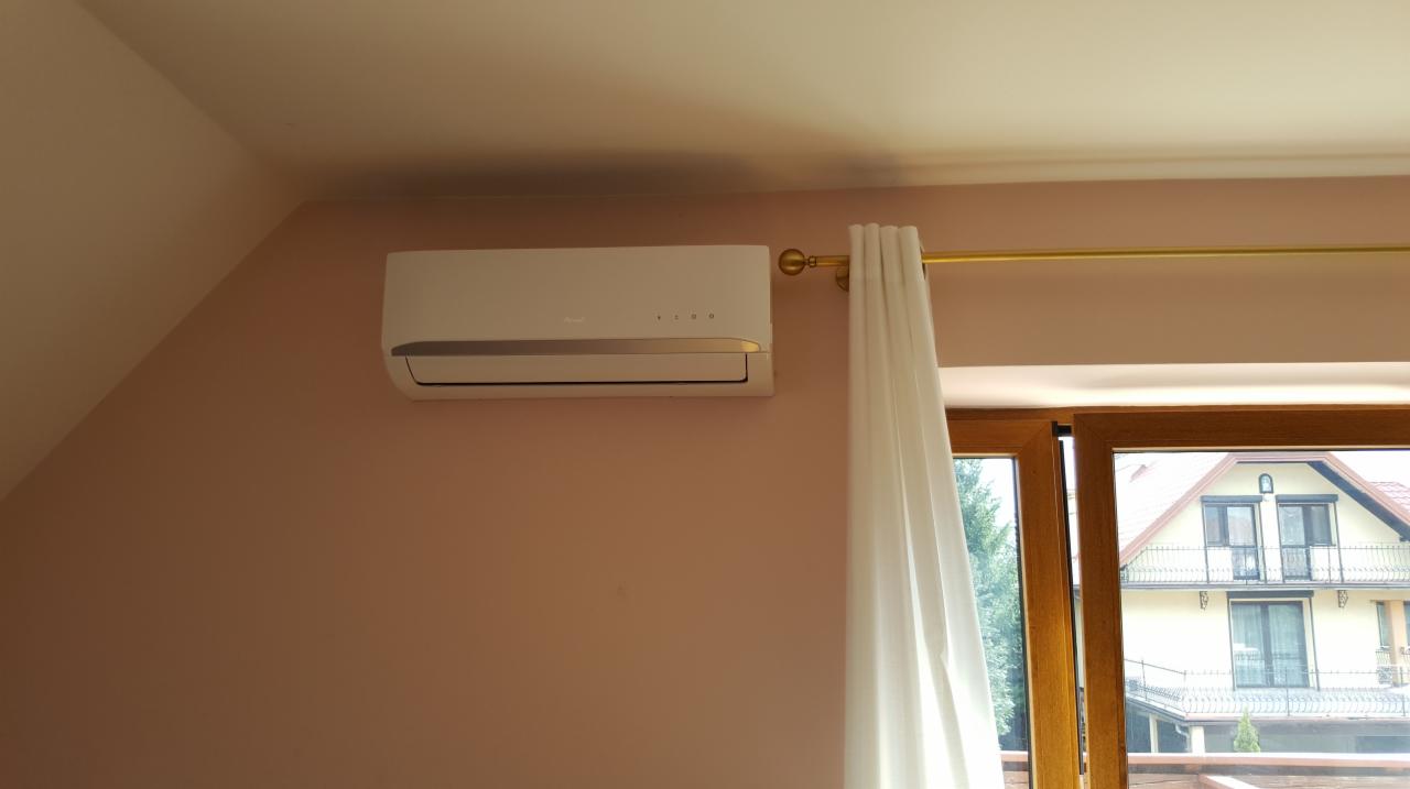 Biały klimatyzator ścienny zamontowany w pokoju z oknem, zasłoniętym białą firanką, widok na sąsiedni dom.