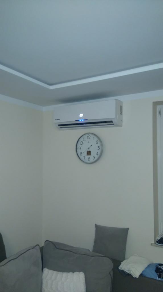 Biała jednostka wewnętrzna zamontowana na ścianie nad okrągłym zegarem w salonie z szarą sofą i dekoracyjną listwą sufitową.