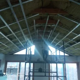 Montaż metalowego rusztu pod płyty na ocieplonym poddaszu z widocznymi oknami, pracownik na rusztowaniu w trakcie pracy.