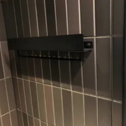 Ściana w łazience wyłożona pionowymi, grafitowymi płytkami ceramicznymi, z zamontowanym czarnym, metalowym wieszakiem na ręczniki.