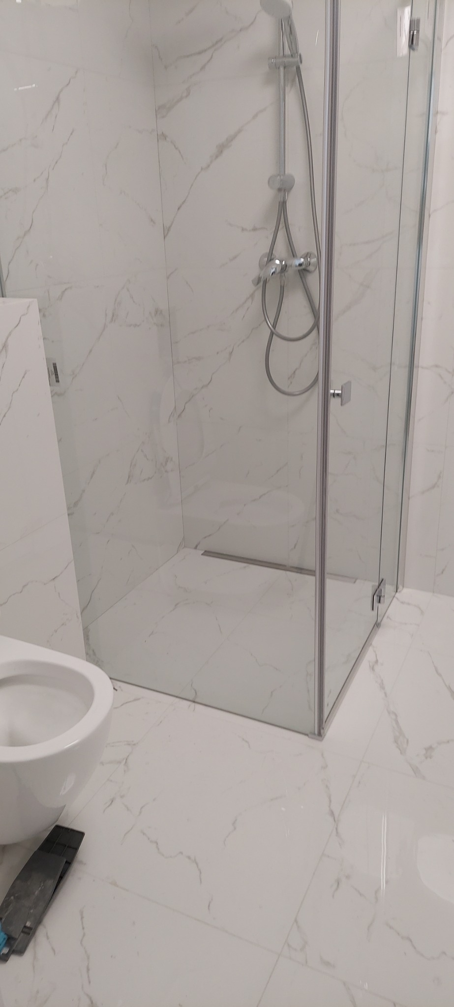 Nowoczesna kabina prysznicowa z chromowaną armaturą i odpływem liniowym, białe płytki imitujące marmur na ścianach i podłodze, widoczny fragment miski WC i narzędzie glazurnicze.