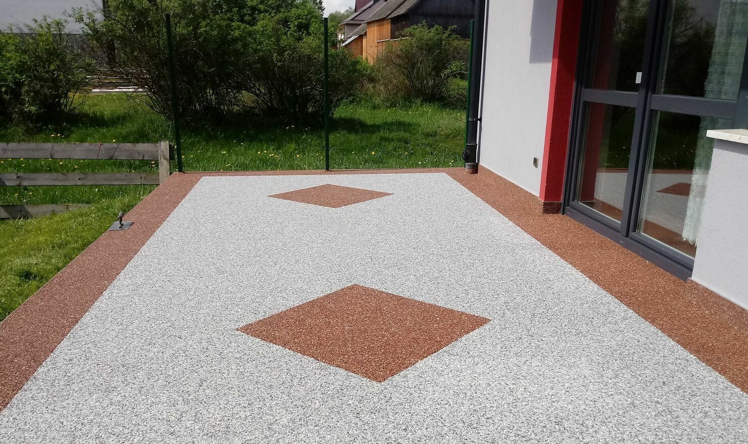 Wykończony taras z żywicy epoksydowej w odcieniach szarości i brązu z wzorem rombów, widok z zewnątrz, drzwi tarasowe przesuwne, trawnik i ogrodzenie w tle.