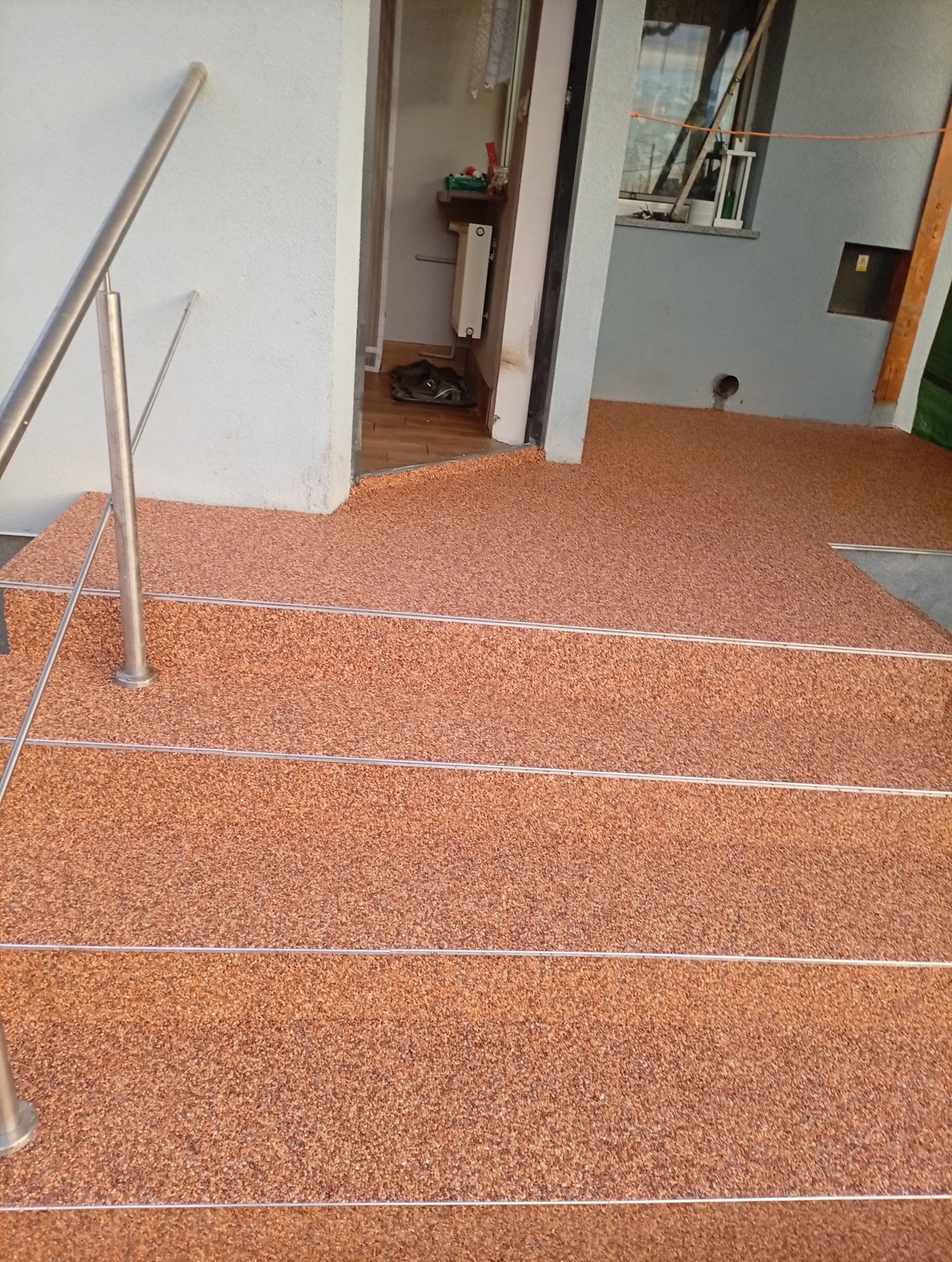 Wykończenie schodów zewnętrznych żywicą epoksydową w kolorze brązowym, z metalowymi listwami antypoślizgowymi. Widok na wejście do budynku.