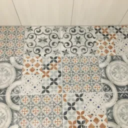 Wzorzyste, patchworkowe płytki podłogowe w odcieniach szarości, błękitu i brązu, ułożone w regularny sposób, kontrastujące z gładką, białą powierzchnią ściany w tle. Widoczne fugi między płytkami.