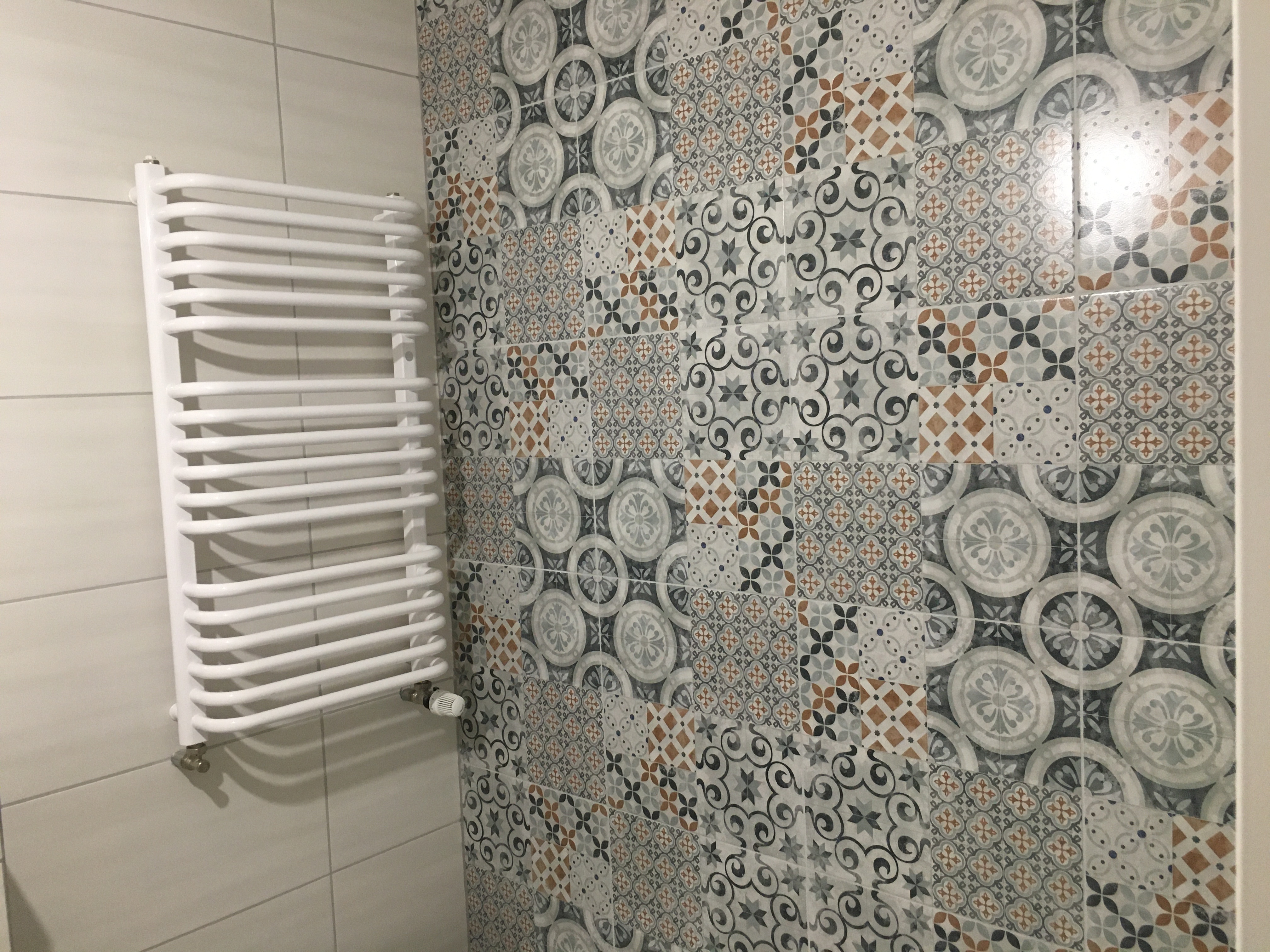Ściana łazienki wyłożona wzorzystymi płytkami w odcieniach szarości, brązu i bieli, z zamontowanym, białym, drabinkowym grzejnikiem. Widoczny zawór termostatyczny na grzejniku.
