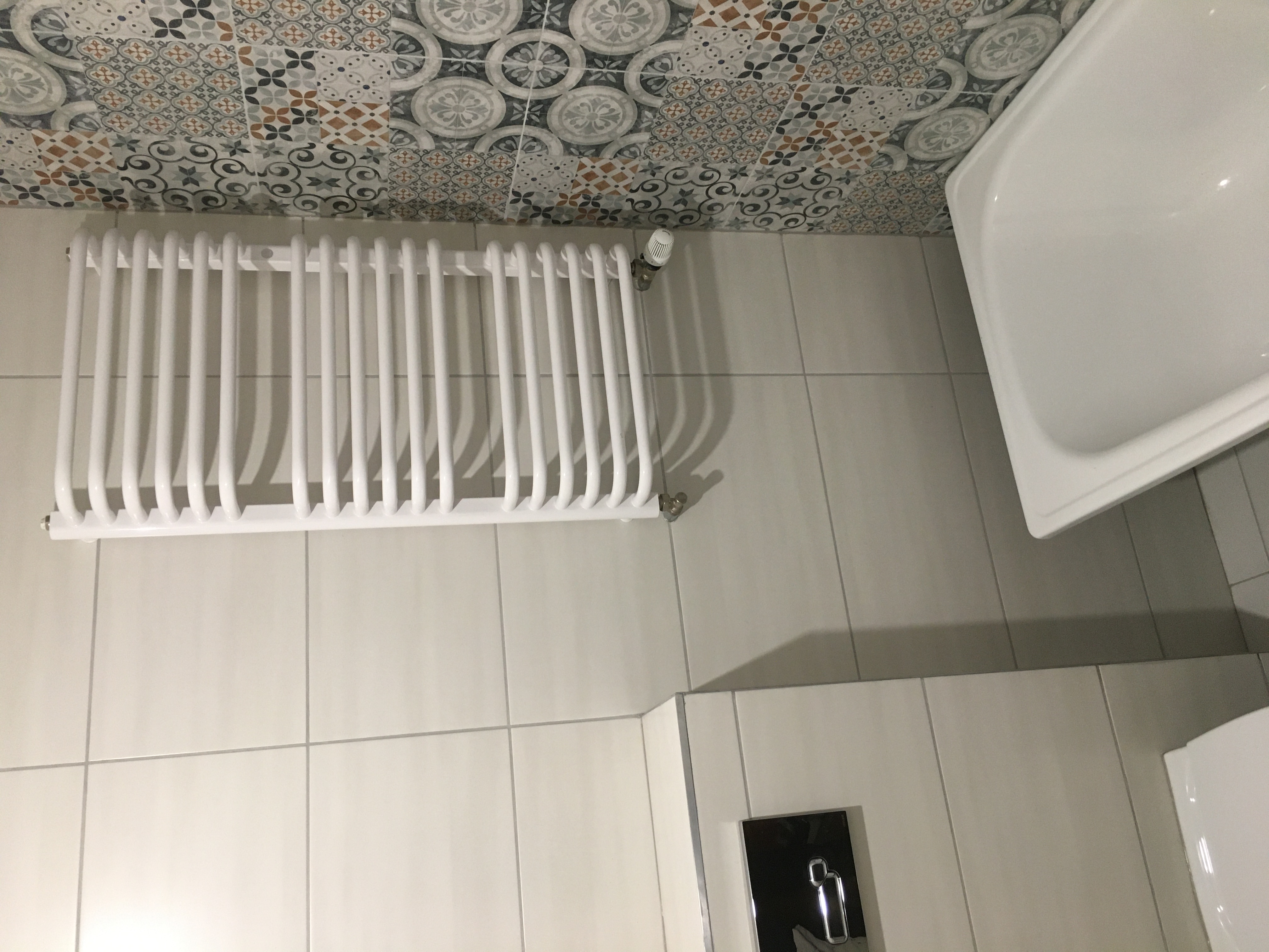 Nowoczesna łazienka z grzejnikiem drabinkowym na tle dekoracyjnych płytek ściennych i jasnych płytek podłogowych, widoczny fragment białej umywalki.