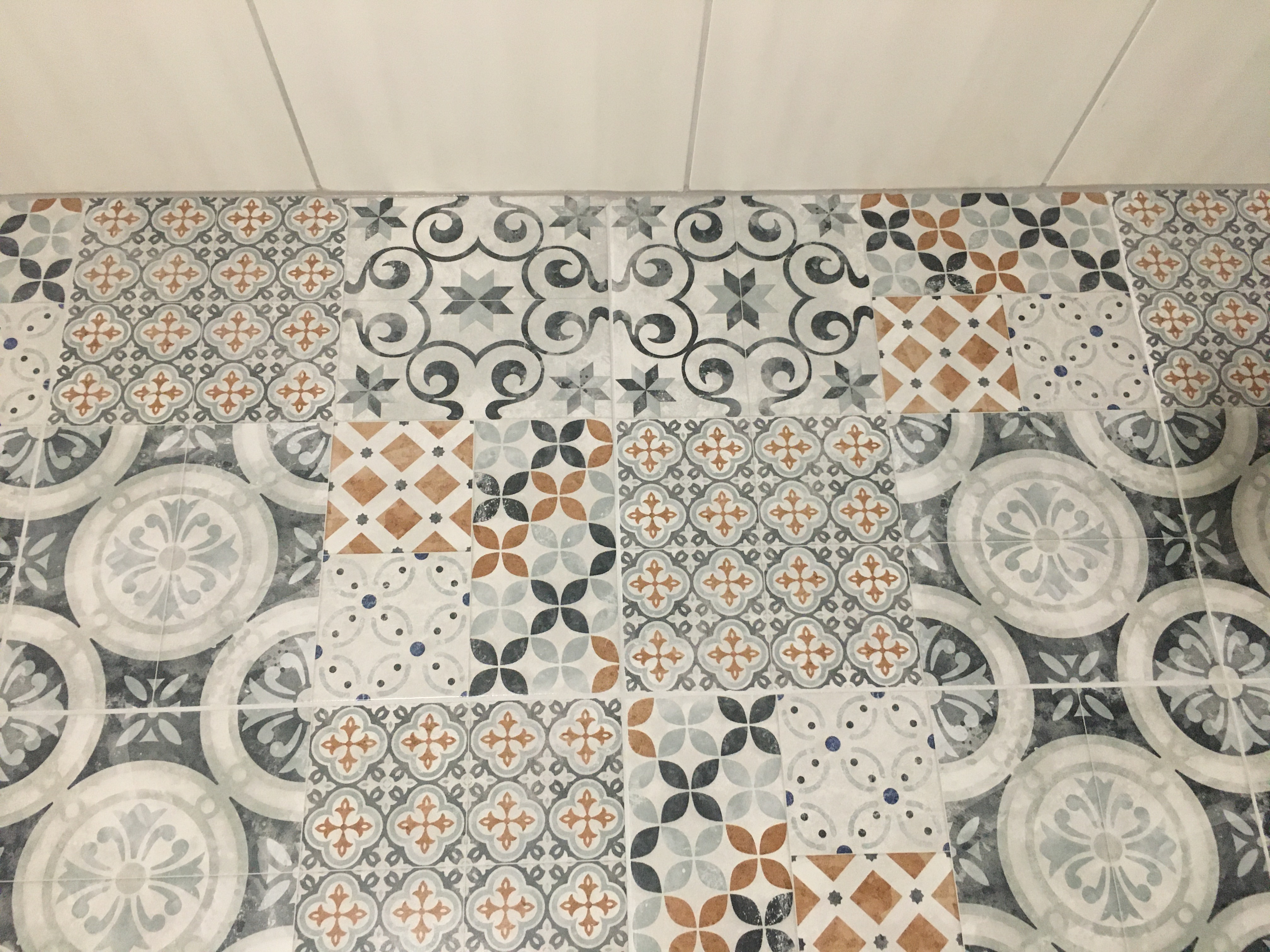 Wzorzyste, patchworkowe płytki podłogowe w odcieniach szarości, błękitu i brązu, ułożone w regularny sposób, kontrastujące z gładką, białą powierzchnią ściany w tle. Widoczne fugi między płytkami.