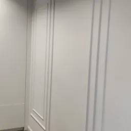 Białe ściany z dekoracyjnymi panelami ściennymi, jasna drewniana podłoga, wentylacja sufitowa.