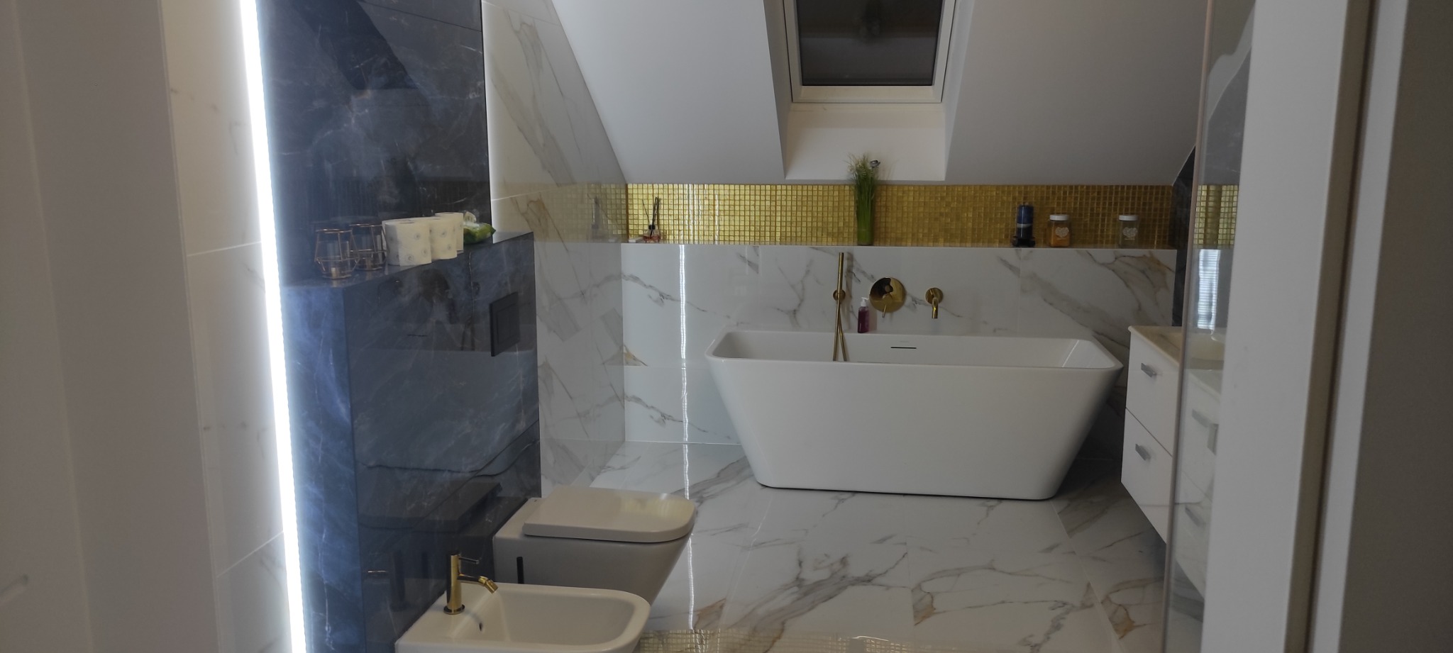 Nowoczesna łazienka na poddaszu z marmurowymi płytkami, wolnostojącą wanną z złotą armaturą, bidetem i toaletą w zabudowie oraz złotą mozaiką na ścianie.