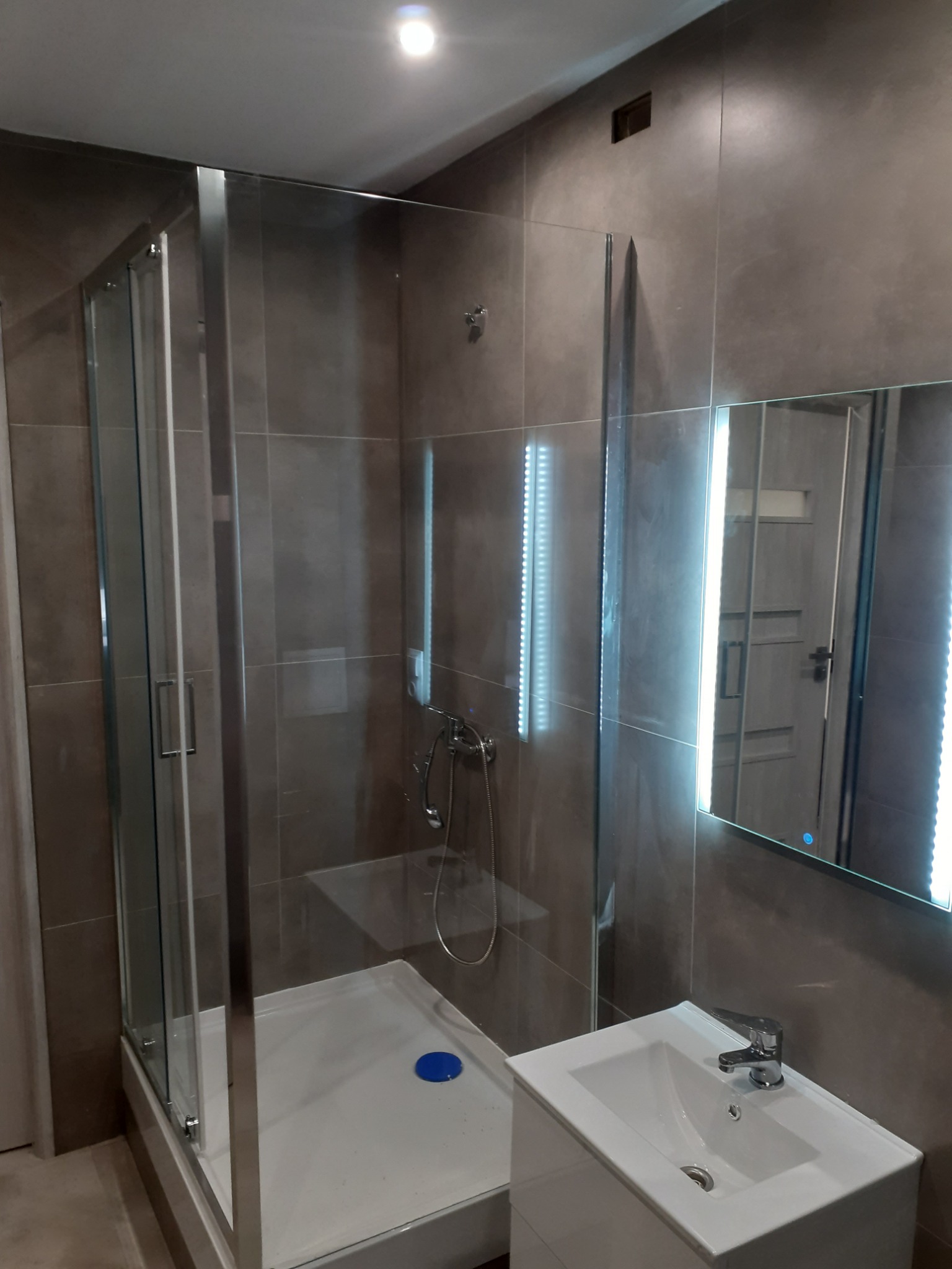 Nowoczesna łazienka po remoncie: kabina prysznicowa z przesuwnymi drzwiami, umywalka z szafką i podświetlane lustro na tle szarych płytek.