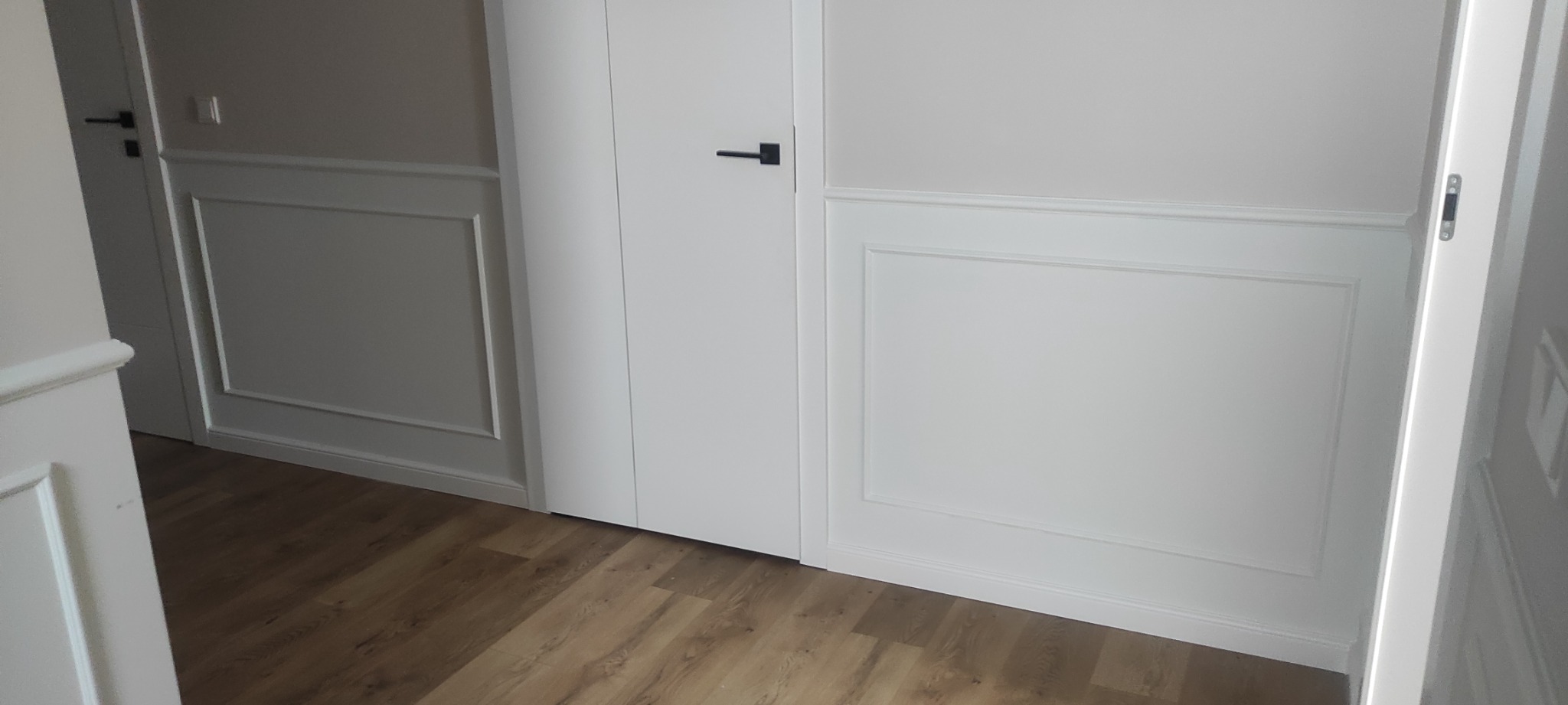 Eleganckie panele ścienne w kolorze białym z subtelnymi listwami dekoracyjnymi, uzupełnione przez drewnopodobną podłogę i minimalistyczne, czarne klamki drzwiowe.