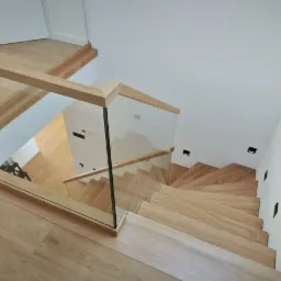 Drewniane schody z balustradą ze szkła i drewna, widok z góry. Parkiet w kolorze naturalnego drewna. Minimalistyczny design z oświetleniem punktowym na ścianie.