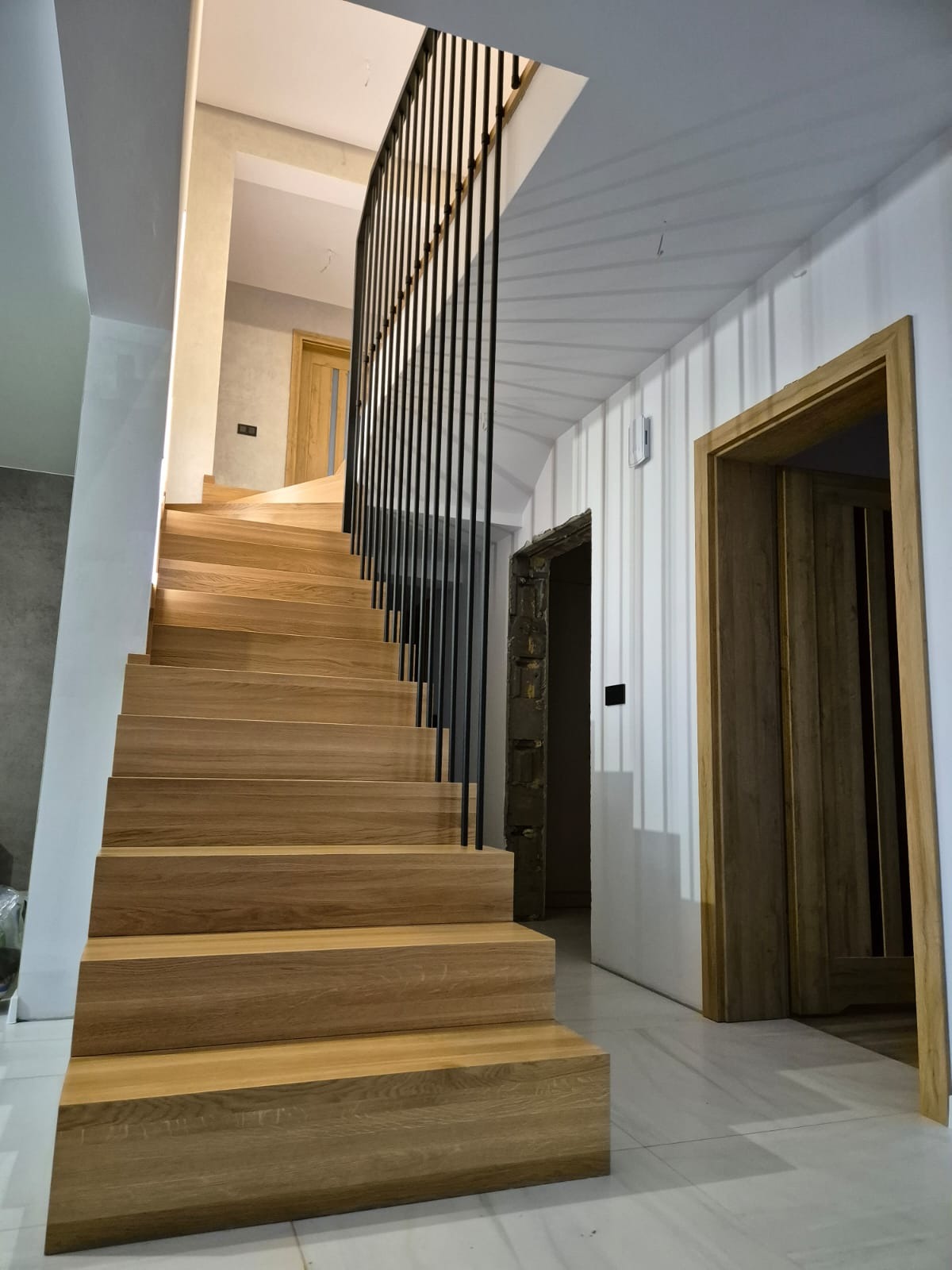 Jasne, drewniane schody z czarną, metalową balustradą w nowoczesnym wnętrzu, widok z dołu, drzwi z drewnianą ościeżnicą w tle.