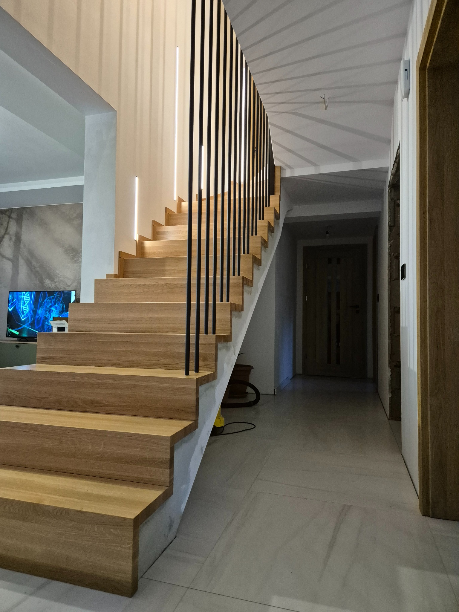 Jasne, dębowe schody z czarną, minimalistyczną balustradą, podświetlane listwami LED w ścianie, prowadzące na piętro. W tle widoczny salon z telewizorem i korytarz z drzwiami.