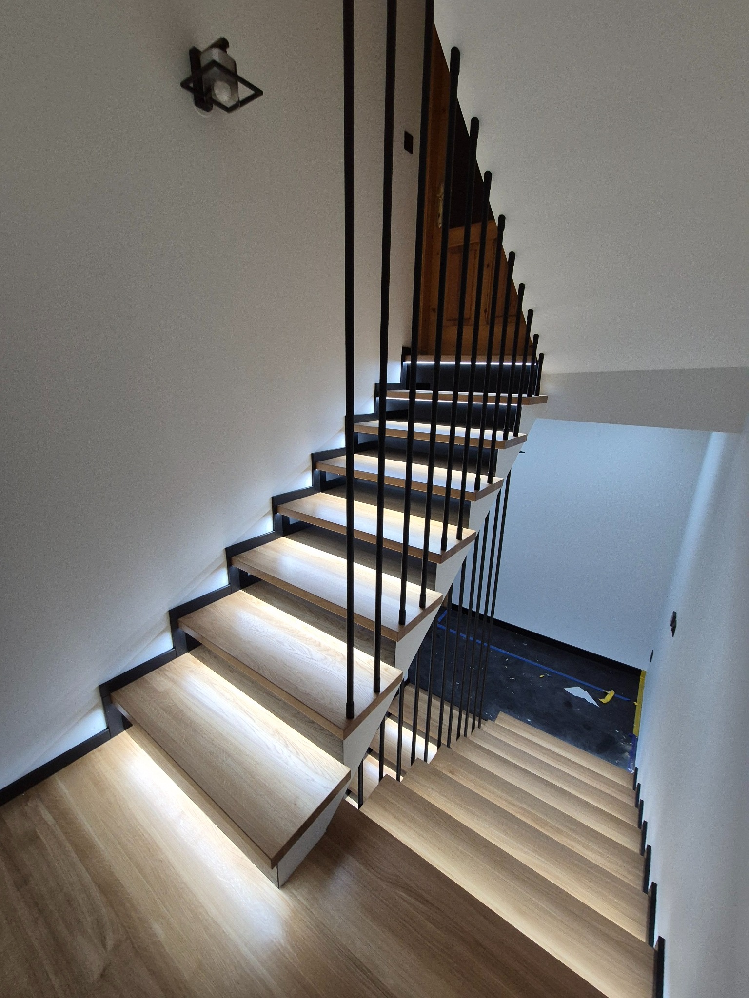 Nowoczesne schody z drewnianymi stopniami podświetlanymi LED, czarne metalowe balustrady pionowe, widok z góry na dół, minimalistyczny design.