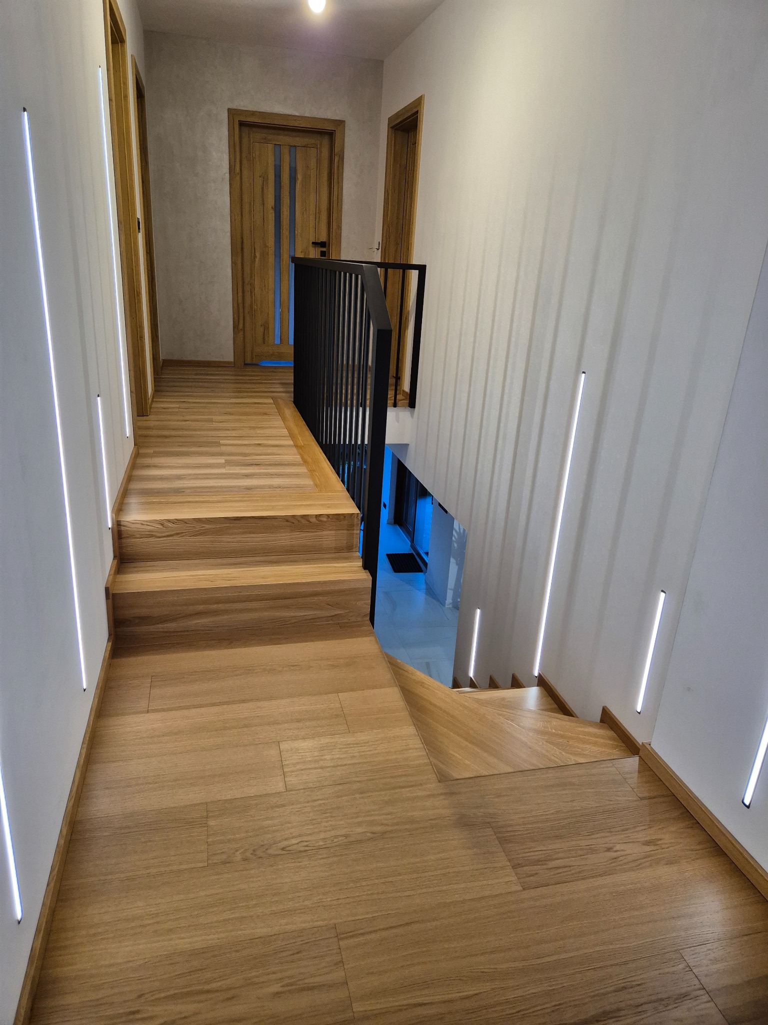 Jasne, drewniane schody z czarną, metalową balustradą, oświetlone pionowymi, ledowymi lampami w ścianie, widok z góry.