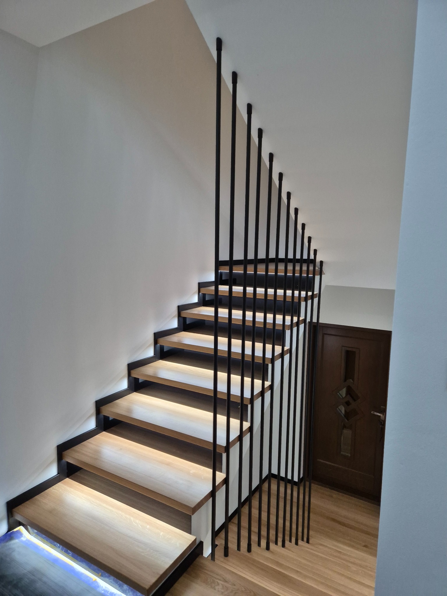 Nowoczesne schody z drewnianymi stopniami, czarną metalową konstrukcją i pionowymi balustradami, oświetlone od spodu, widok z góry.