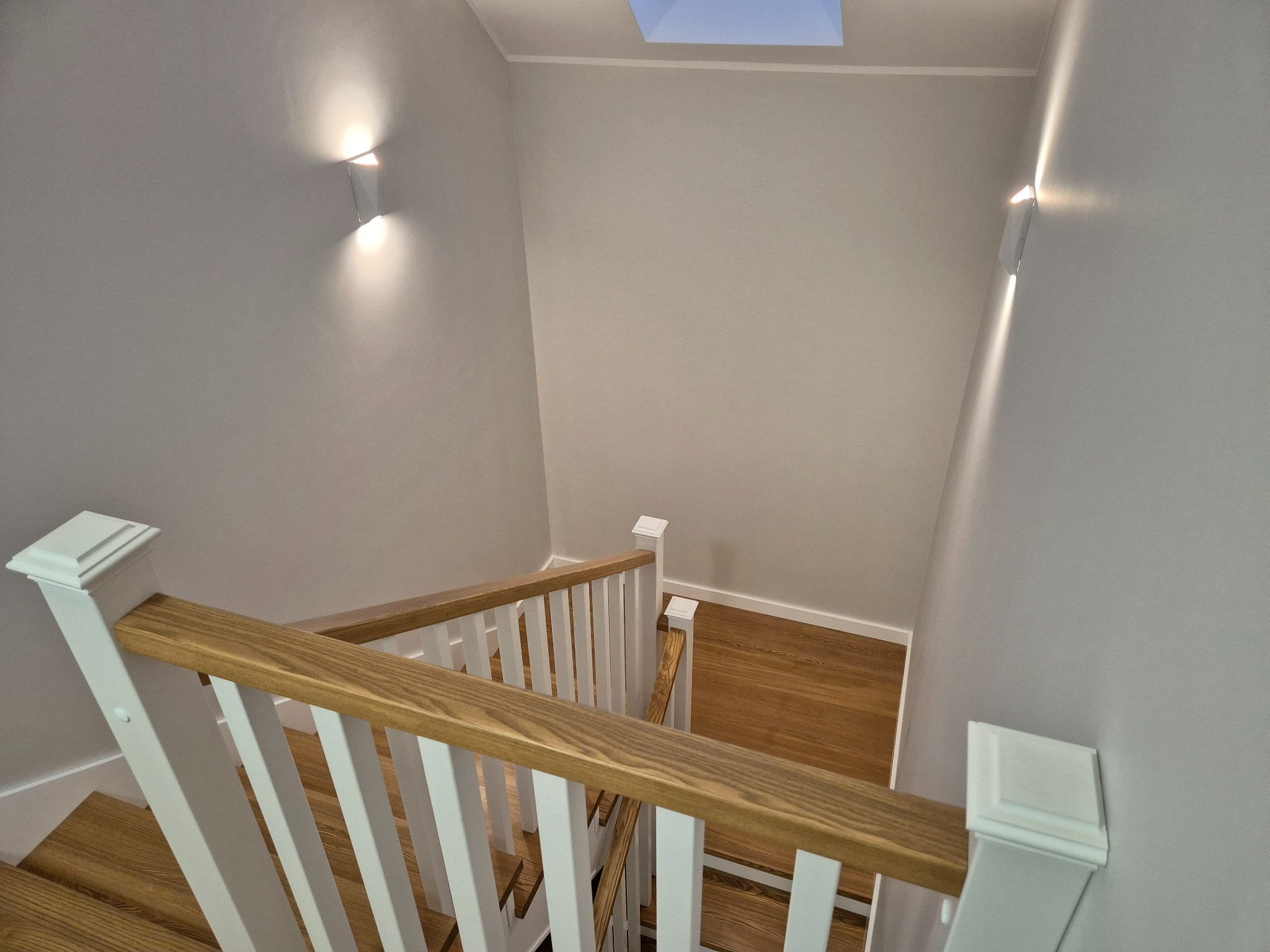 Widok z góry na drewniane schody z białymi balustradami i oświetleniem ściennym, z widocznym świetlikiem dachowym.