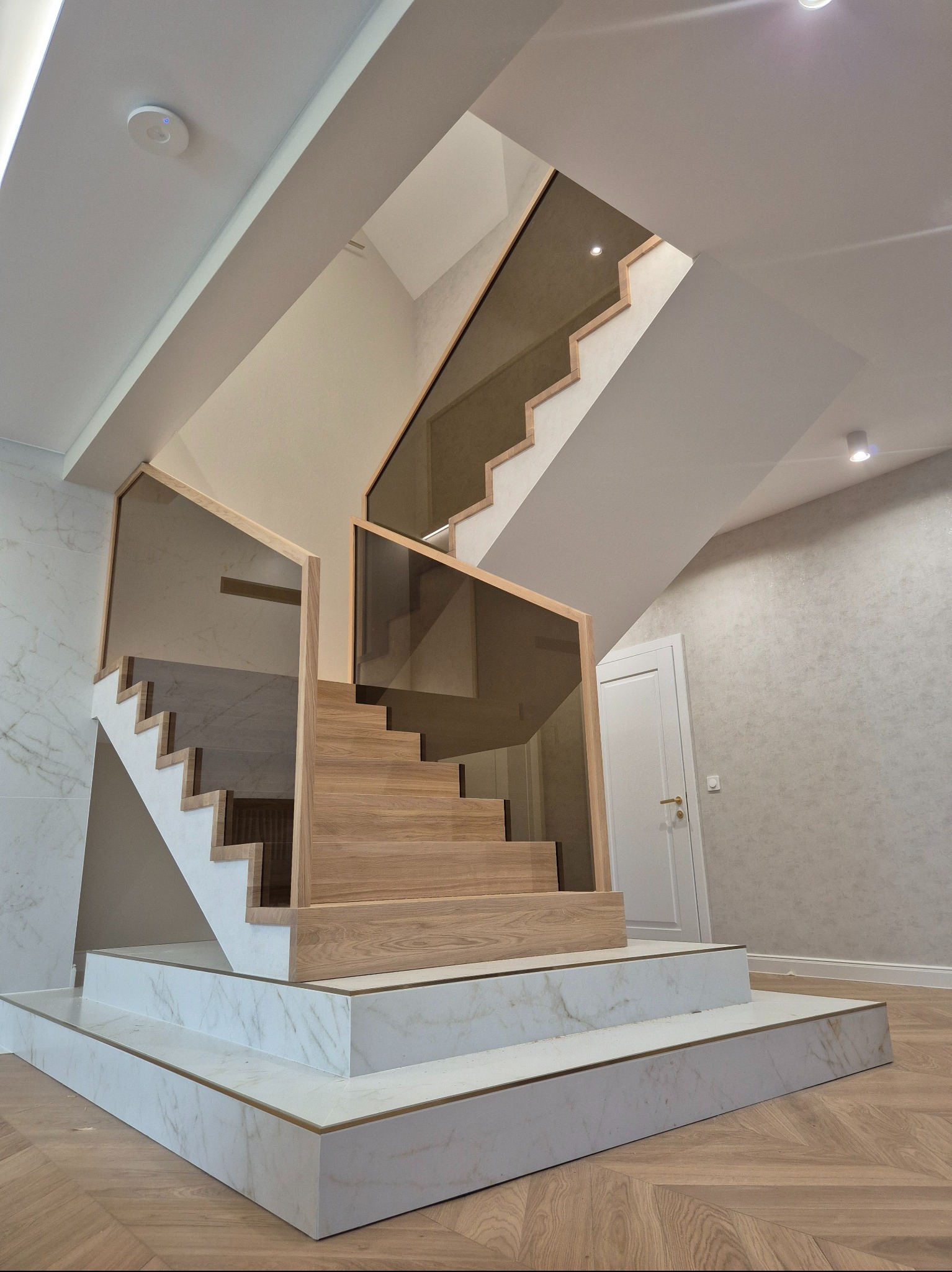 Nowoczesne schody z drewnianymi stopniami i szklaną balustradą, wykończone marmurem, w jasnym wnętrzu z parkietem w jodełkę i białą stolarką.