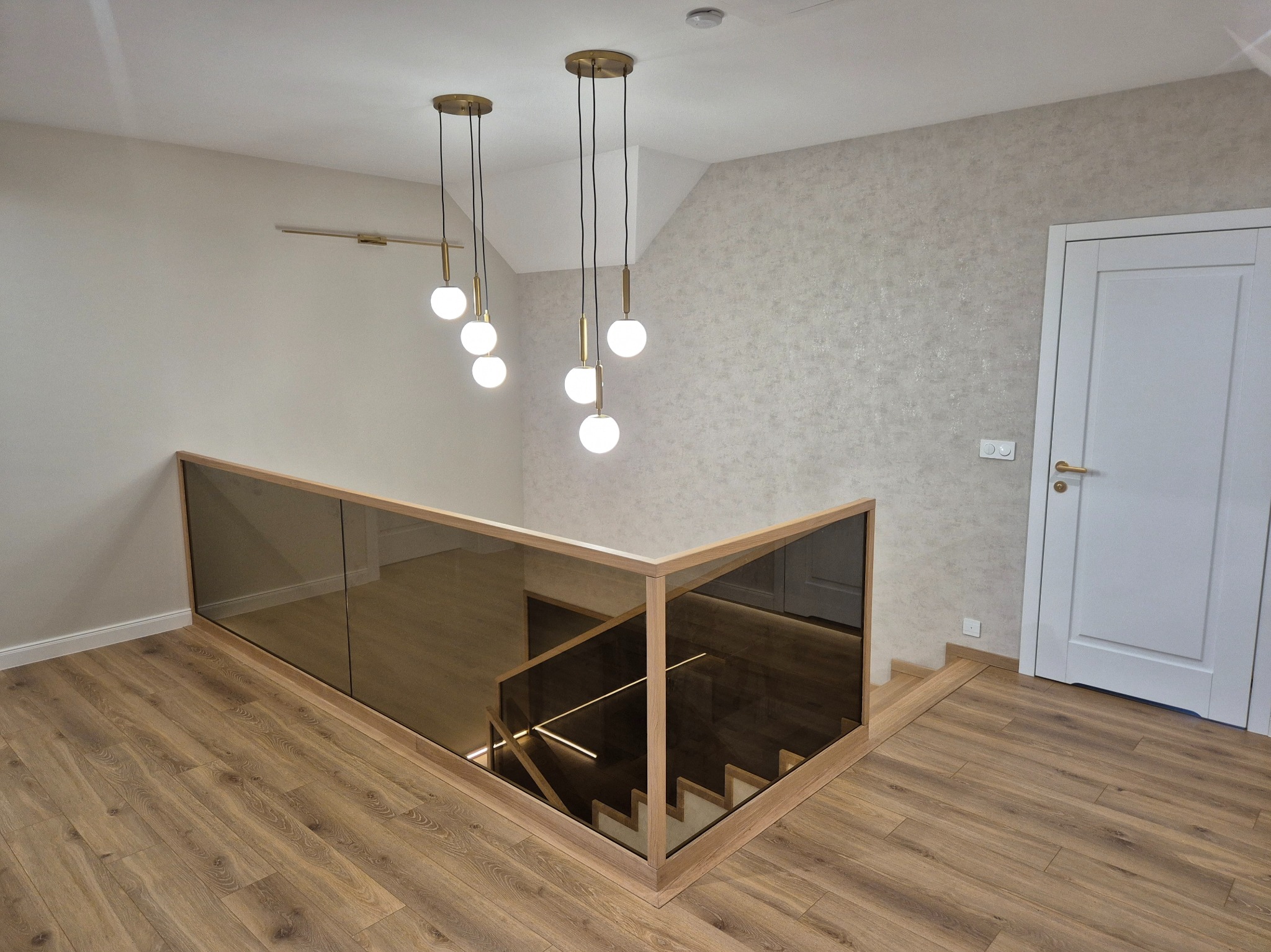 Nowoczesne schody z drewnianą ramą i szklaną balustradą. Oświetlenie LED na schodach i designerskie lampy wiszące. Parkiet w ciepłym odcieniu drewna i jasna ściana.