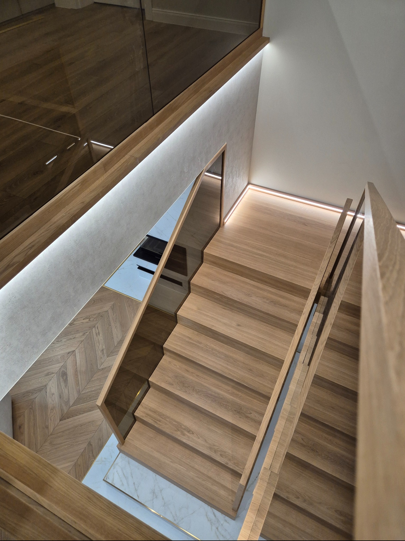 Nowoczesne schody drewniane z podświetleniem LED i szklaną balustradą, widok z góry, parkiet w jodełkę w tle. Jasne drewno i minimalistyczny design.