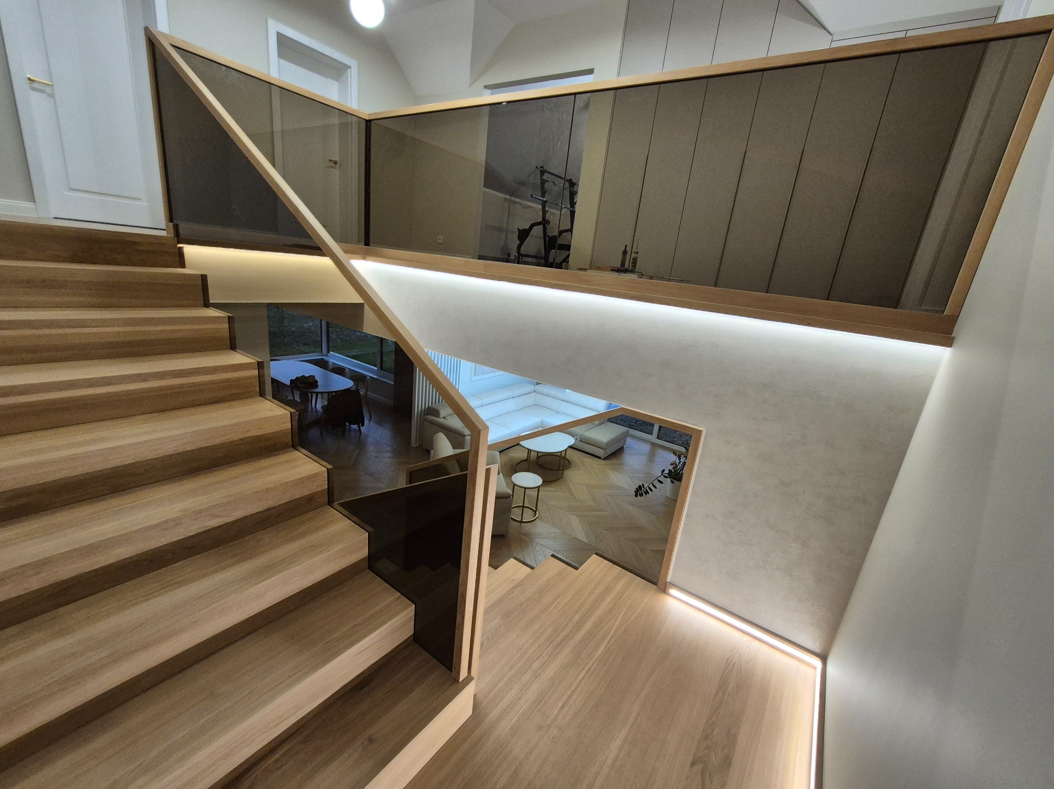 Nowoczesne schody z drewnianymi stopniami i szklaną balustradą z drewnianą ramą, oświetlone LED, w domu w Hucisku. Widok na salon z góry.