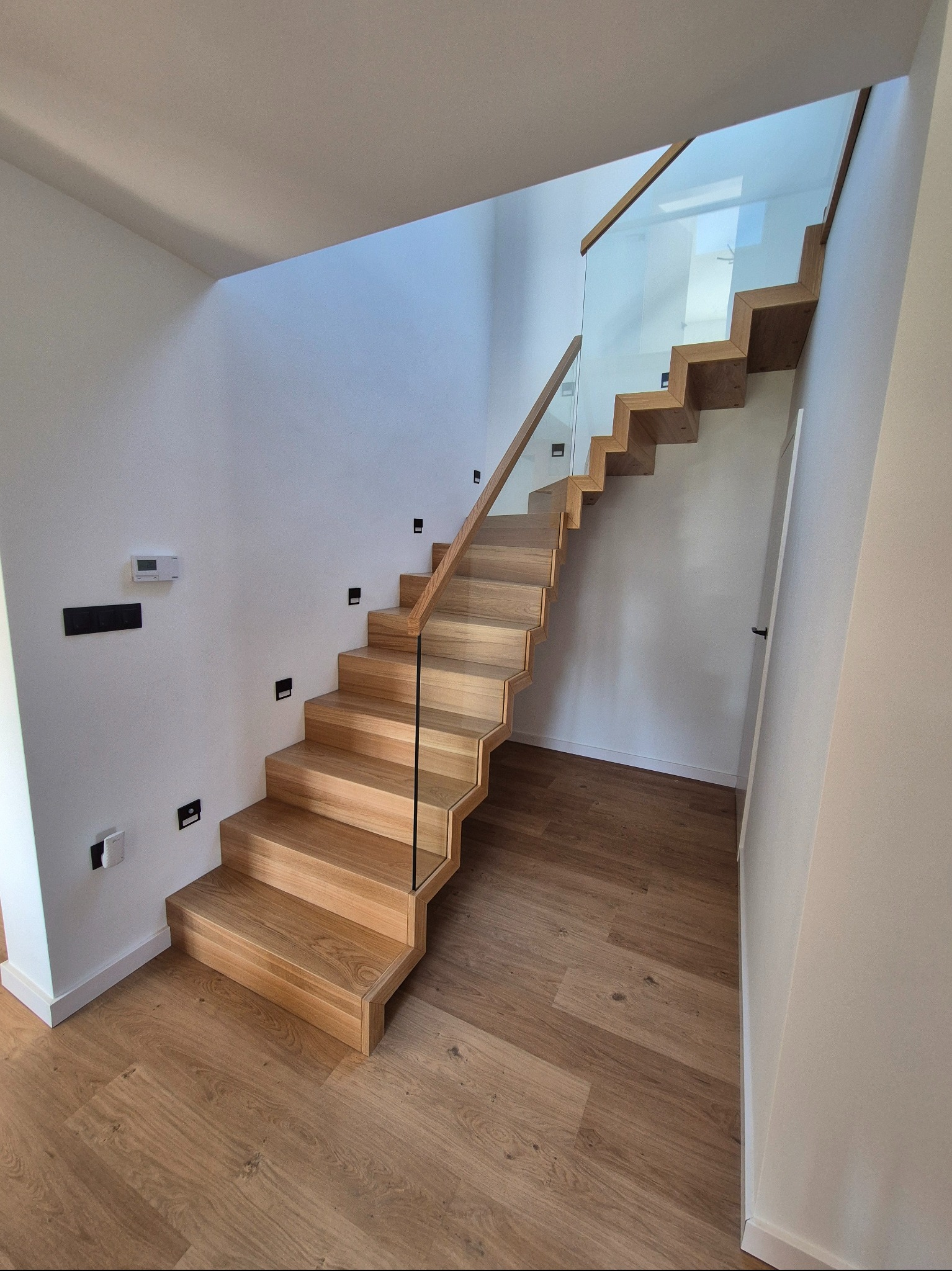 Nowoczesne schody z jasnego drewna z szklaną balustradą, oświetleniem punktowym na ścianie, w minimalistycznym wnętrzu. Widok z dołu, z dbałością o detale wykończenia.