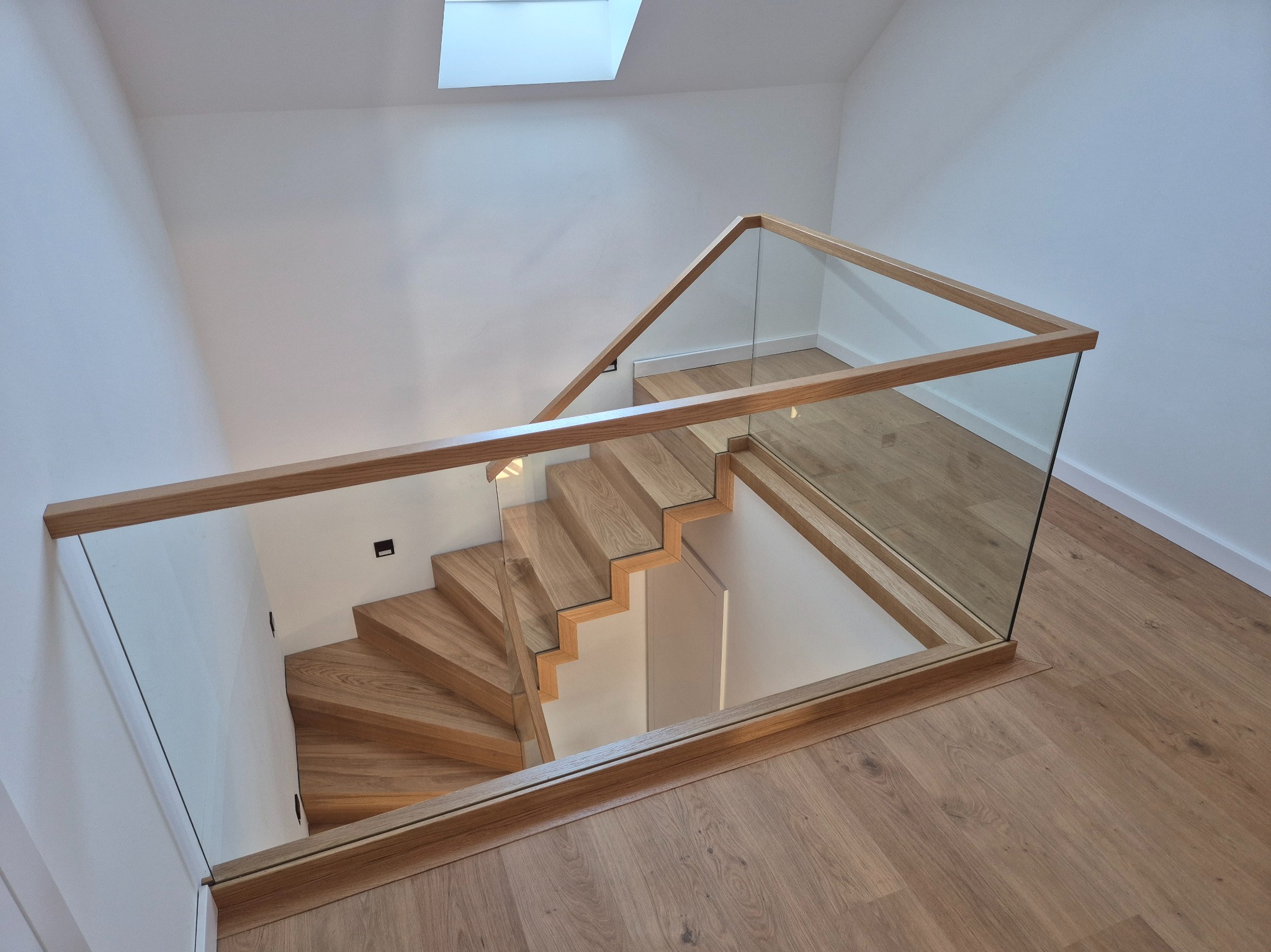 Kątowe ujęcie na drewniane schody z przeszkloną balustradą w jasnym wnętrzu. Widoczne drewniane stopnie i podłoga, białe ściany i sufit z oknem dachowym.