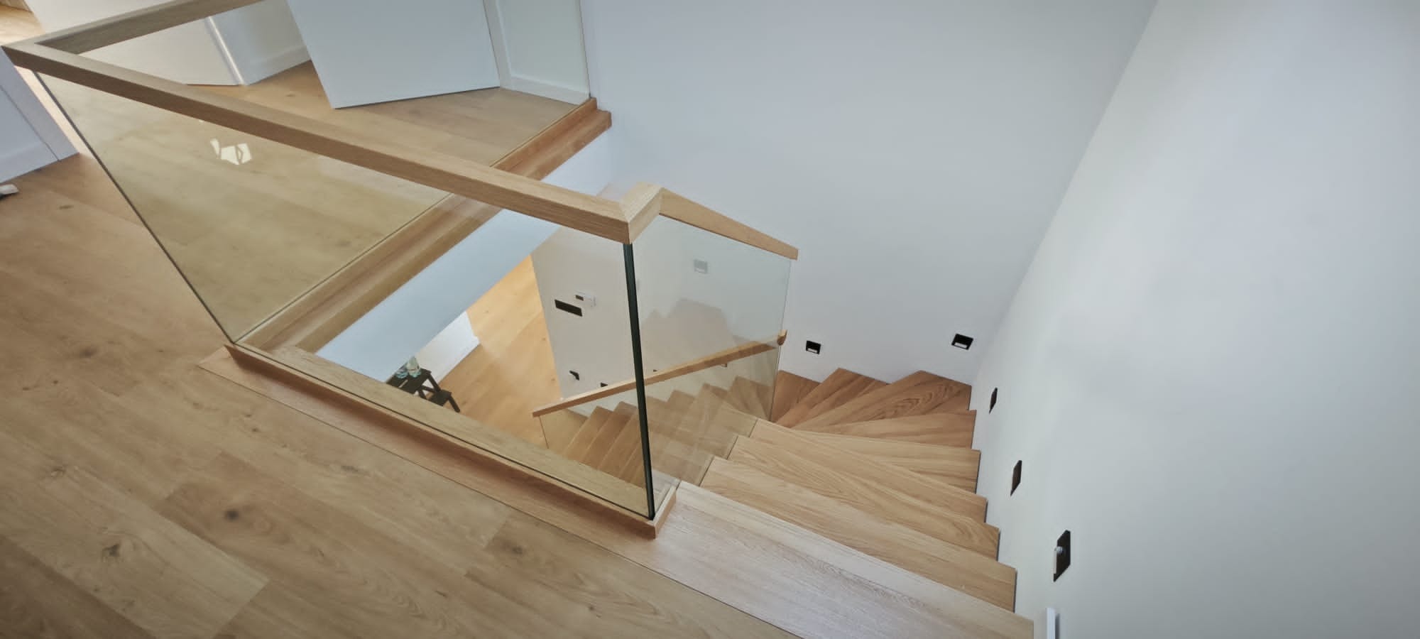 Drewniane schody z balustradą ze szkła i drewna, widok z góry. Parkiet w kolorze naturalnego drewna. Minimalistyczny design z oświetleniem punktowym na ścianie.