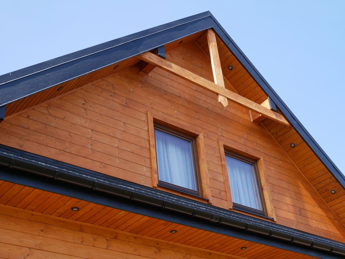 Fragment ściany szczytowej domu z drewnianą elewacją, dwoma oknami z białymi zasłonami i czarnym dachem oraz rynnami na tle błękitnego nieba.