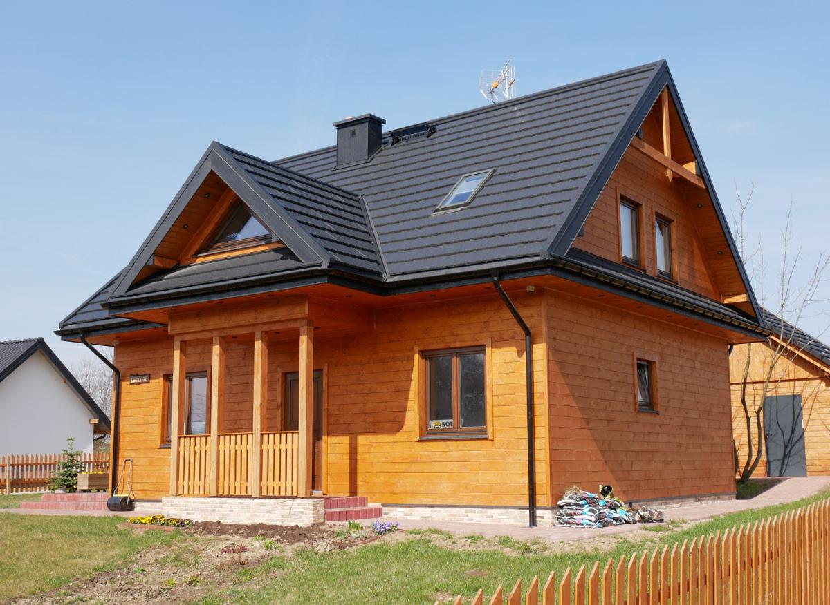 Gotowy, parterowy budynek z drewna z gankiem, ciemną dachówką i anteną satelitarną na dachu, widok z przodu.