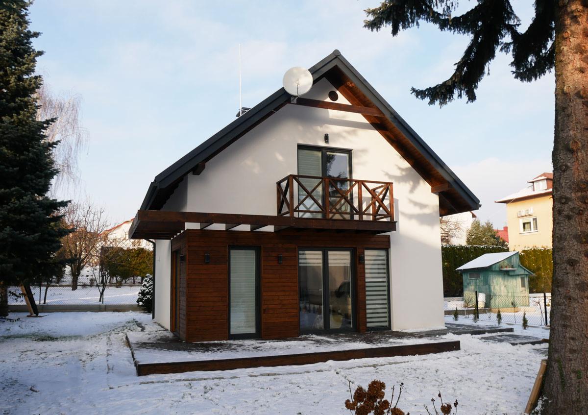 Nowoczesny dom z białymi ścianami i drewnianą elewacją, balkonem z drewnianą balustradą i anteną satelitarną na dachu, otoczony ośnieżonym ogrodem.