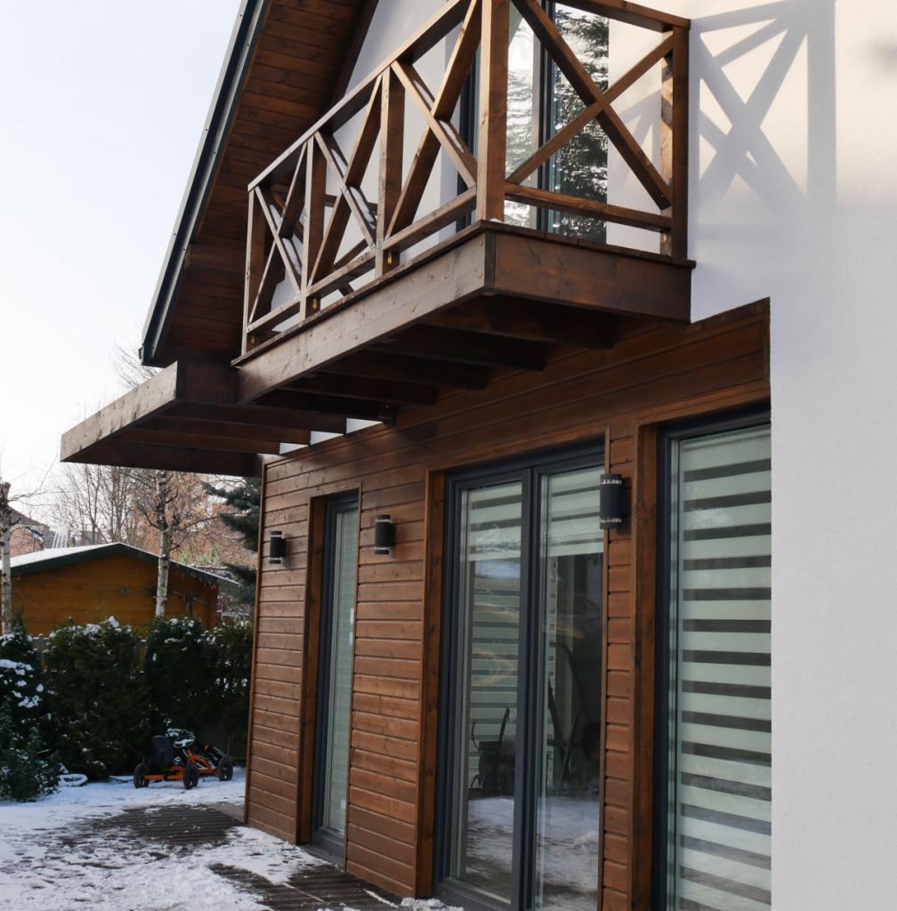 Fragment domu szkieletowego z drewnianą elewacją, balkonem z balustradą w kształcie krzyży i dużymi oknami przesuwnymi z żaluzjami w zimowej scenerii.