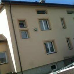 JackBud - Trzykondygnacyjny budynek po ociepleniu i pomalowaniu elewacji na jasny beż, widoczne białe ramy okienne, ciemny pas pod dachem i fragment niebieskiego nieba, ujęcie z dołu.