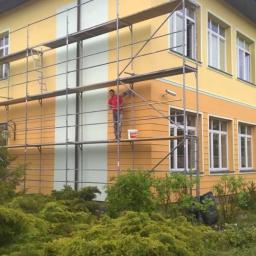 JackBud - Pracownik na rusztowaniu maluje fragment elewacji budynku na jasnozielony kolor, kontrastujący z żółtą fasadą. Widoczny wiadro z farbą i otaczająca zieleń.