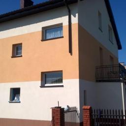 JackBud - Dwukondygnacyjny dom z elewacją w odcieniach brązu, beżu i bieli, widoczne okna z firankami i balkon z czarną, kutą balustradą, fragment czerwonego samochodu w rogu kadru.