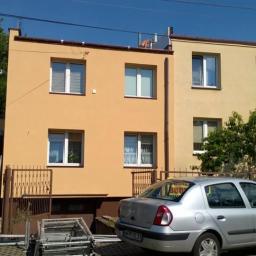 JackBud - Dwukondygnacyjny dom z pomarańczową i żółtą elewacją, częściowo odnowiony, z widocznym pracownikiem na drabinie malującym fragment ściany, srebrny samochód zaparkowany przed budynkiem.