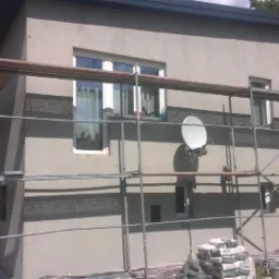 Dom jednorodzinny w trakcie tynkowania elewacji, widoczne rusztowania, siatka pod tynk i worki z zaprawą. Na ścianie zamontowana antena satelitarna.