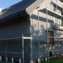 Dom jednorodzinny w trakcie ocieplania szarymi płytami styropianowymi, widoczne rusztowania i fragmenty niezabezpieczonej elewacji.