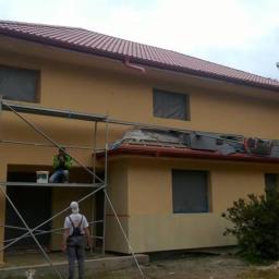 JackBud - Dwupiętrowy dom w trakcie malowania elewacji na żółto, widoczne rusztowania, pracownicy, okna zabezpieczone folią, fragment pomalowanej dolnej części ściany w jaśniejszym odcieniu.